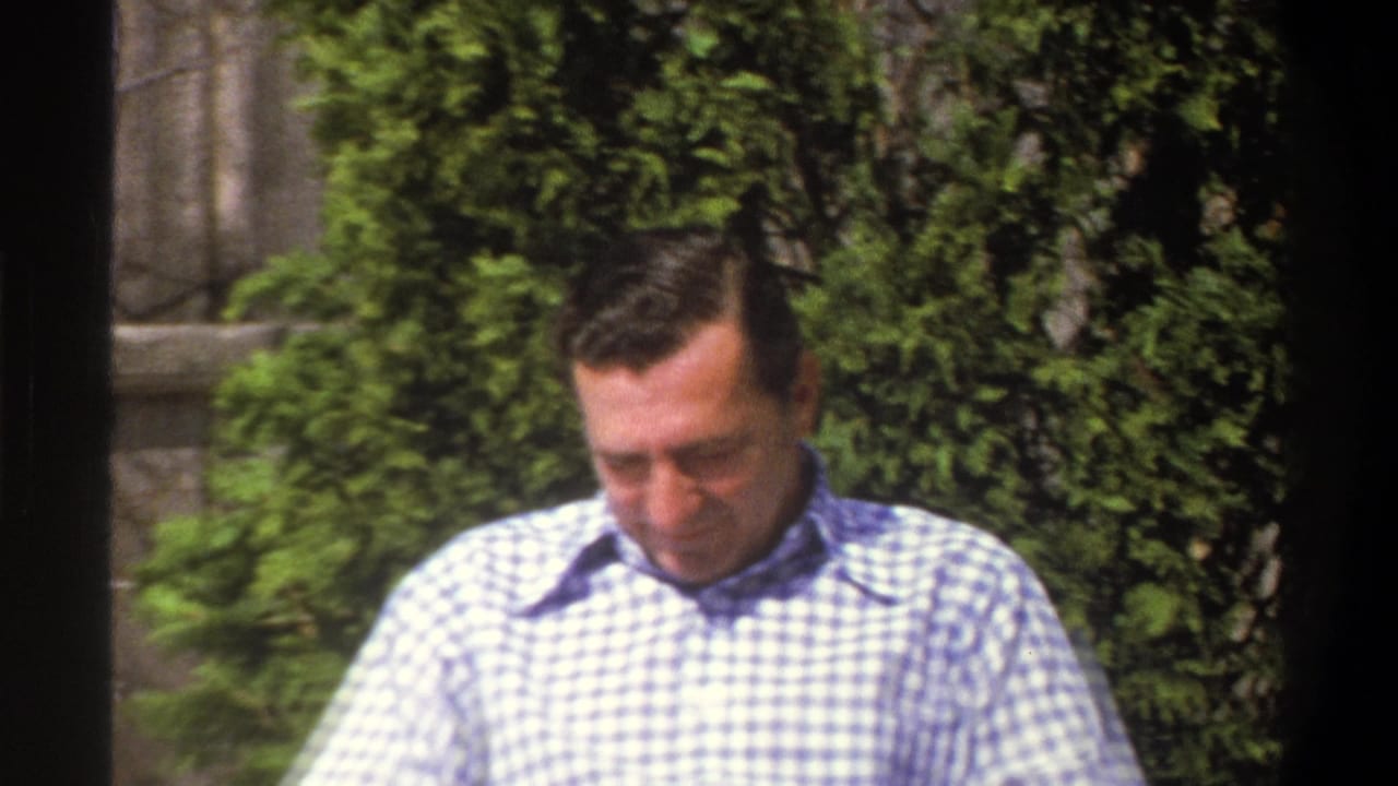 A man in a blue and white checkered shirt stands outdoors near dense greenery in metadata-supported 1969 Cape Town footage.