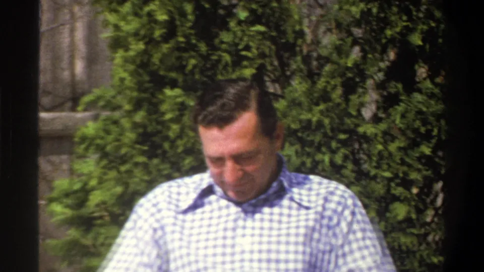 A man in a blue and white checkered shirt stands outdoors near dense greenery in metadata-supported 1969 Cape Town footage.