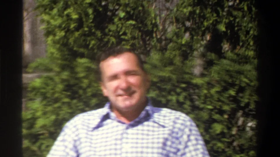 The man smiles toward the camera in a blue and white checkered shirt, framed by green shrubs in 1969 Cape Town home footage.