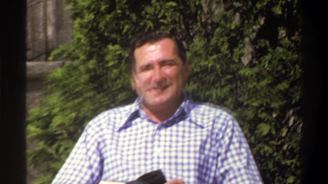 In 1969 Cape Town home-movie footage, an adult man in a blue and white checkered shirt sits outdoors and smiles toward the camera.