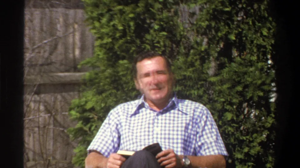 A wider 1969 Cape Town frame shows the man seated outdoors, holding a small item near his lap before a hedge and fence.