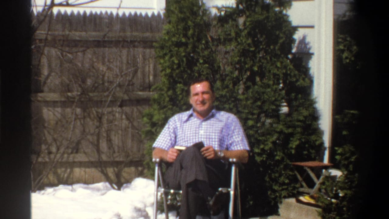 Man seated in a metal chair in a snowy backyard near shrubs and a wooden fence.