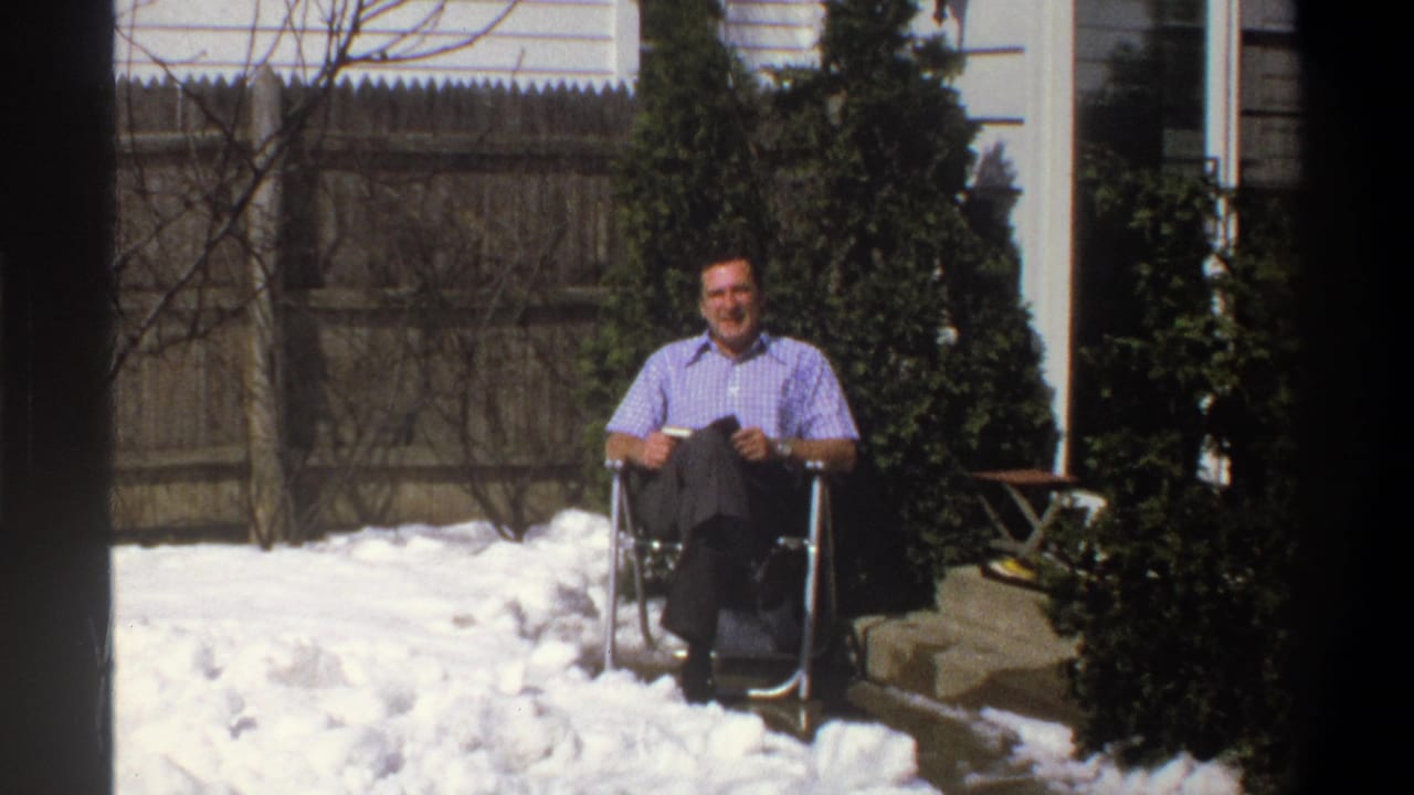 1969 Cape Town frame from the zoom-out, with the seated man smaller in view amid snow, shrubs, fence, and house exterior.