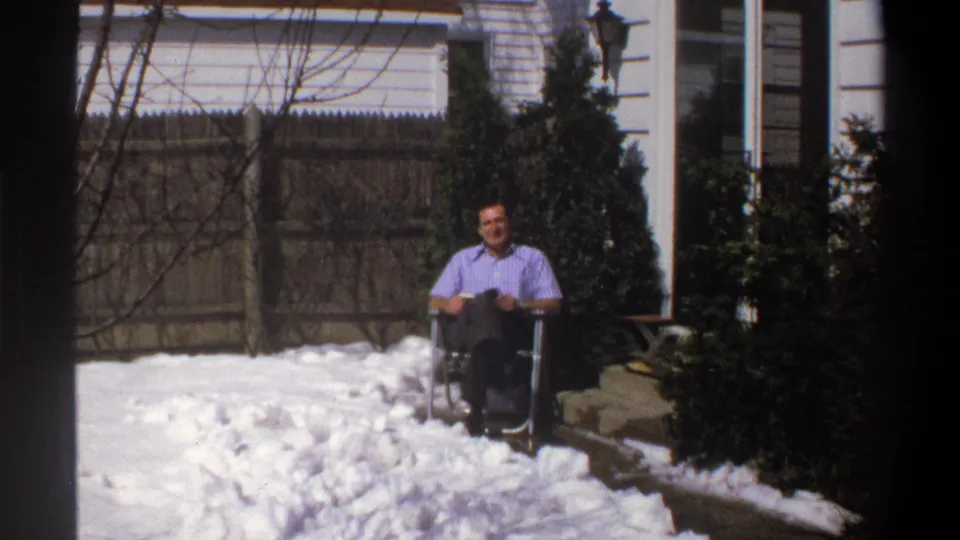 Wider 1969 Cape Town home-movie view of the man seated in a snowy backyard near a fence, shrubs, and part of a house.
