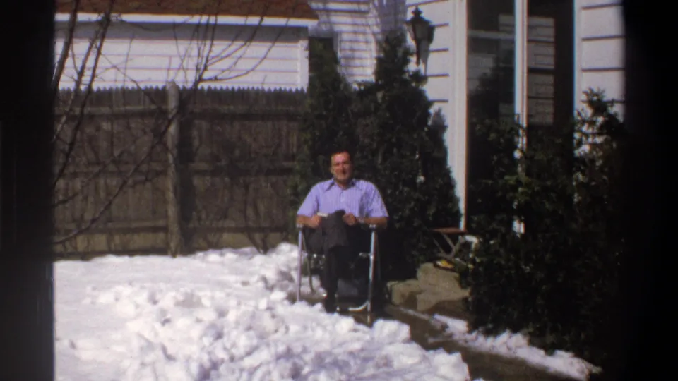 End of the zoom-out shows the man seated near the right side of a snowy backyard, with fence, shrubs, and house exterior visible.