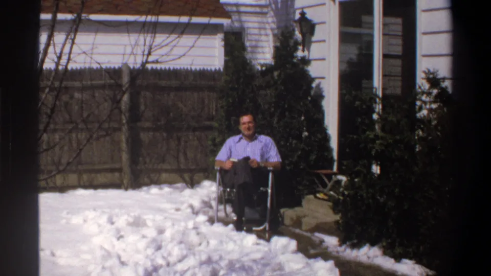 1969 home-movie frame from the supplied Cape Town clip showing a man in a wheelchair facing the camera in a snowy yard.