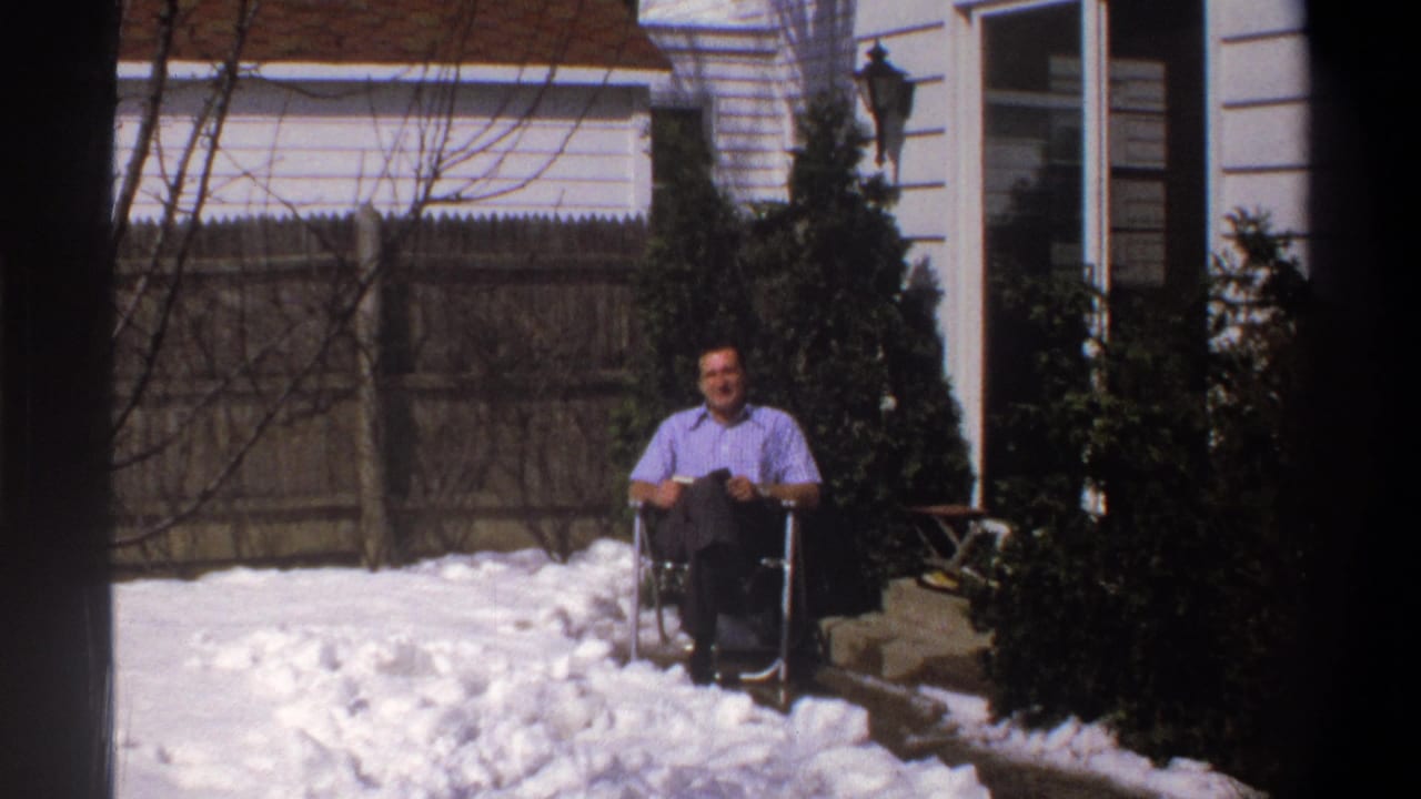 Man in wheelchair smiles toward the camera beside snow and shrubs.