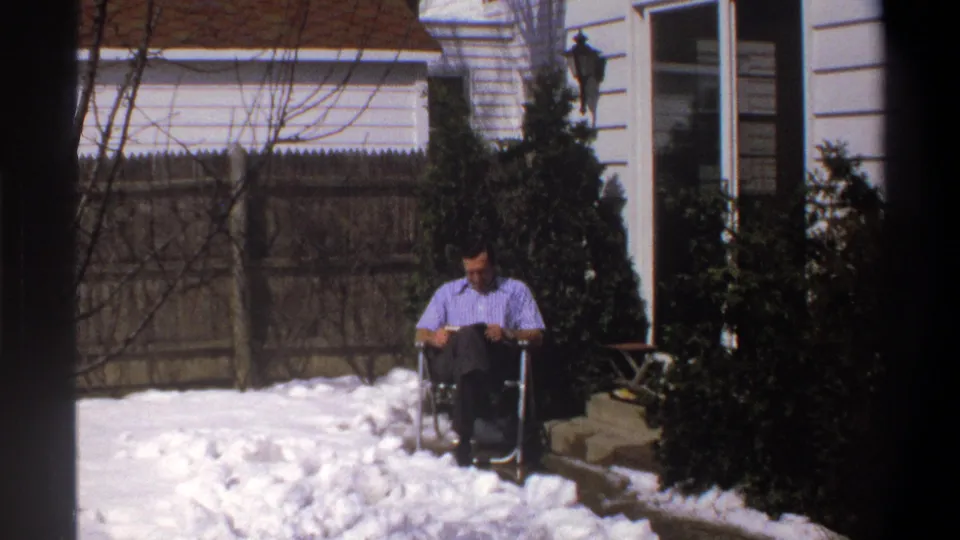 In the supplied 1969 home-movie context, the man lowers his gaze to read while seated in the wheelchair near the house.