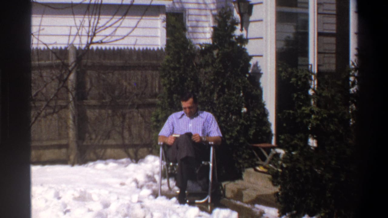 Man seated in a folding chair reading a small book in a snowy yard.