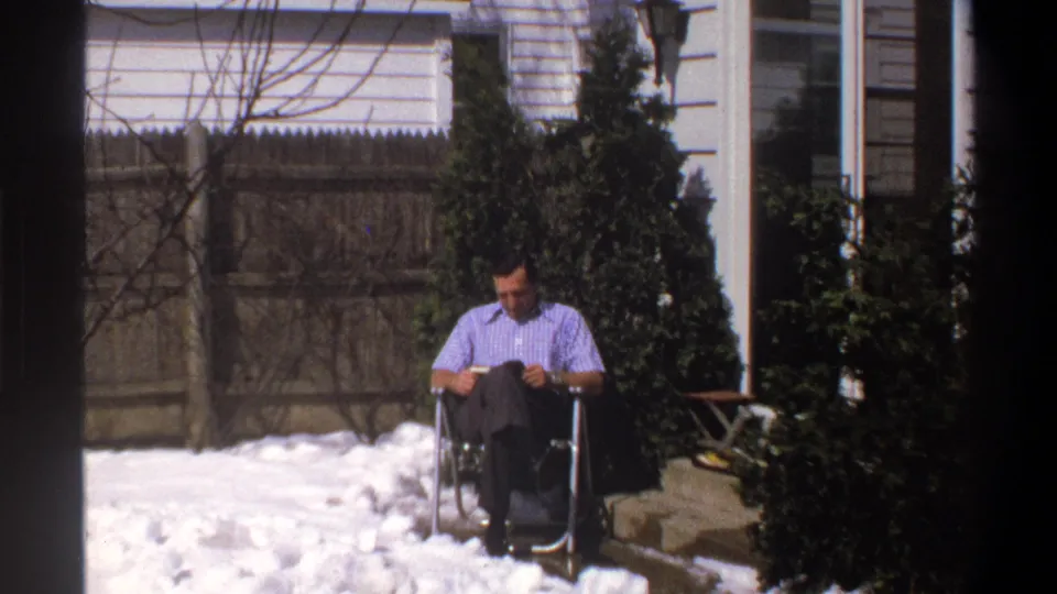1969 Cape Town home-movie frame of a man in a checkered shirt reading outdoors beside snow, shrubs, a fence, and house siding.