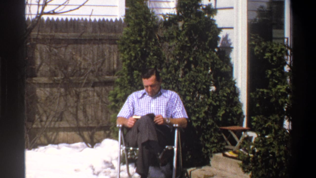 1969 Cape Town home-movie frame showing a seated man reading in bright daylight, with snow, evergreen shrubs, and a wooden fence nearby.