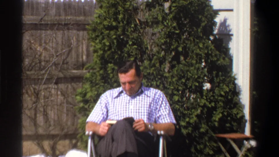 Closer 1969 Cape Town home-movie frame of the man reading in a folding chair, with snow, shrubs, fence boards, and house trim visible.