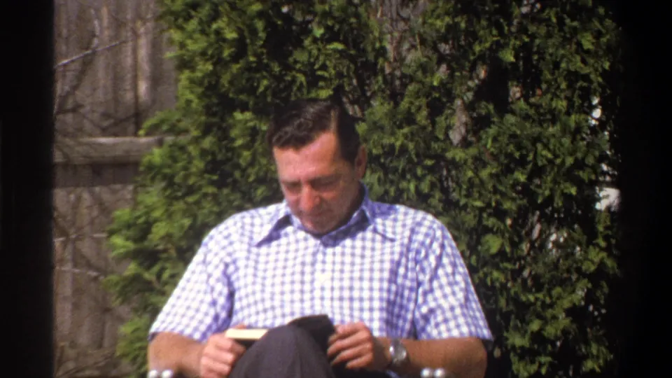 Tight 1969 Cape Town home-movie frame of the seated man holding a small book, with evergreen shrubs and a wooden fence behind him.
