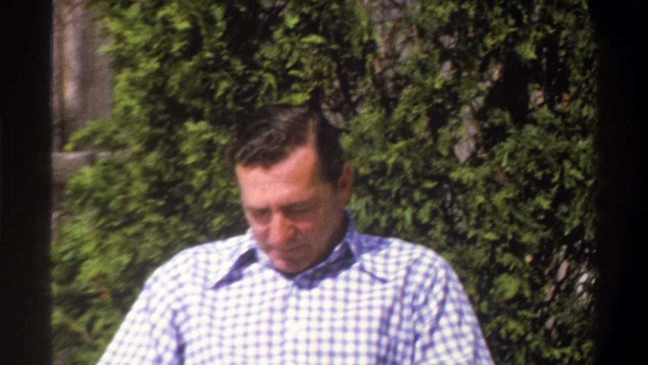 Seated man in a checkered shirt looking down outdoors beside green foliage.