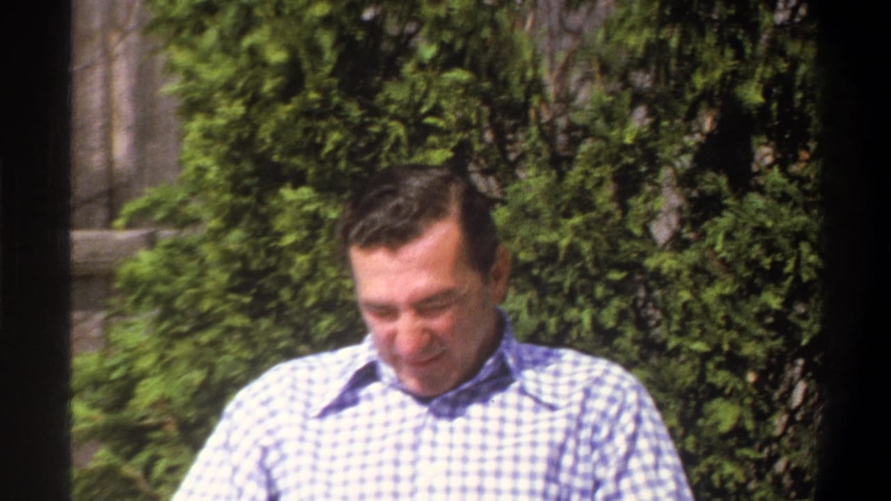 The seated man begins to smile in the Cape Town 1969 home-movie scene, with dense greenery behind him.