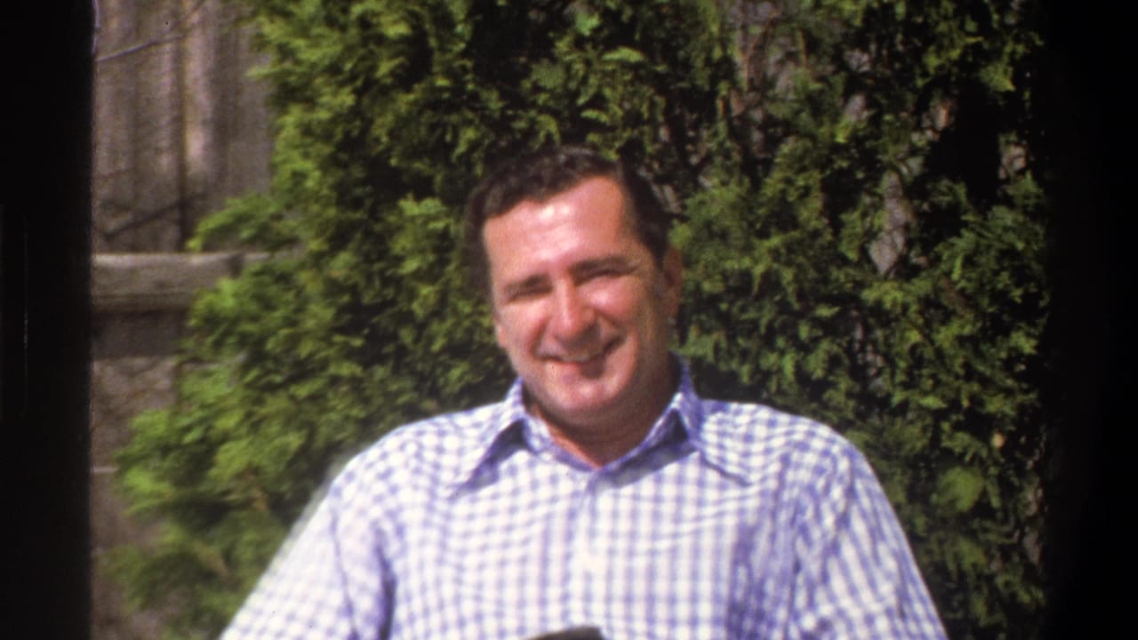 Smiling adult man in a checked shirt seated outdoors before foliage and a stone wall.
