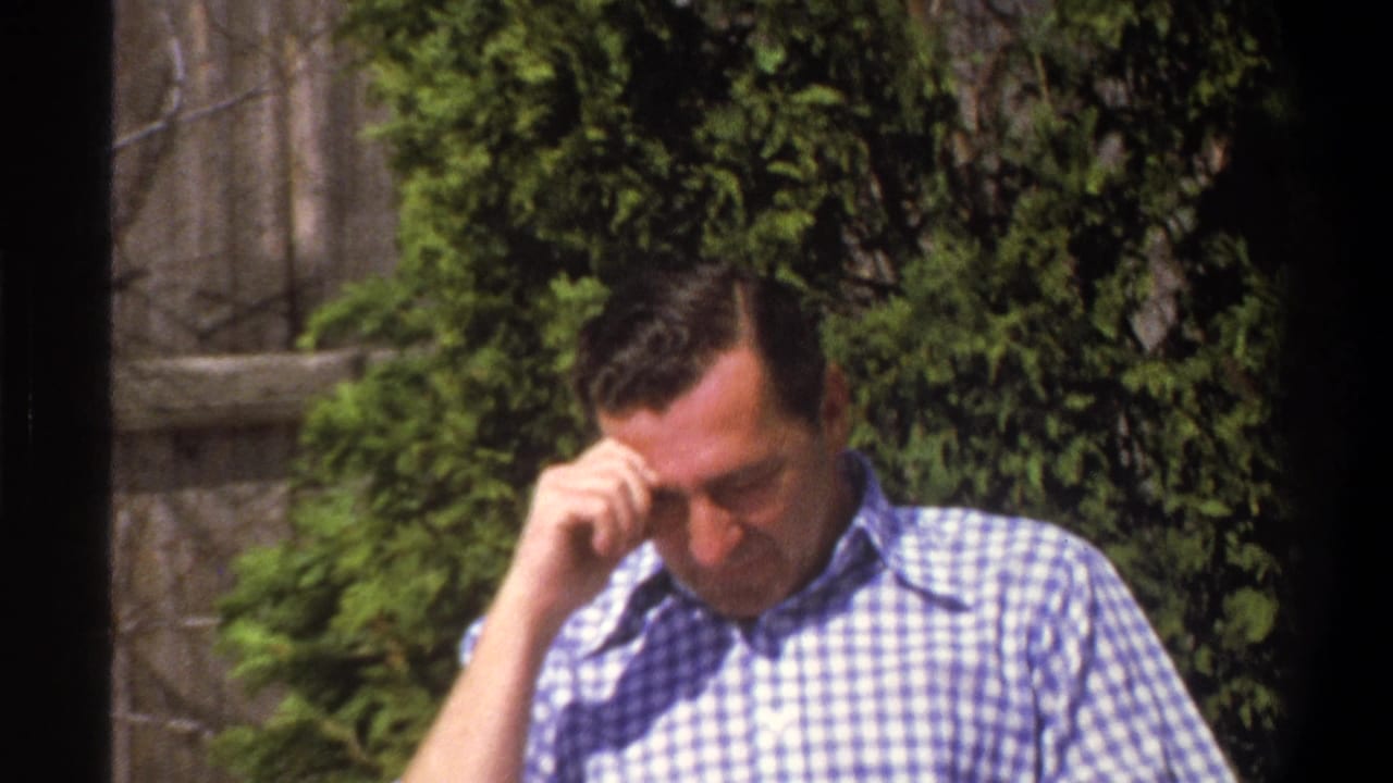 Adult man in checked shirt looks down with one hand near his forehead outdoors.