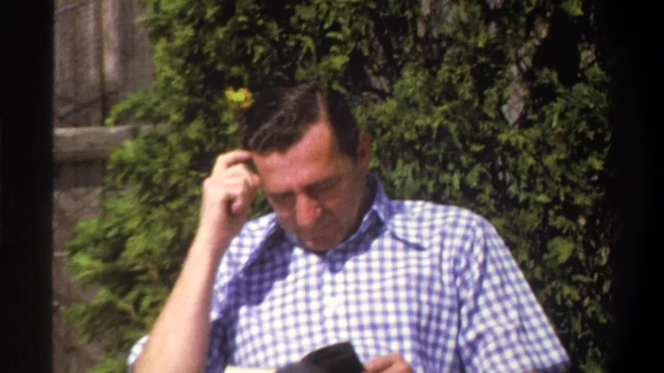 A 1969 Cape Town home-movie frame shows the man touching his forehead and looking down toward a dark book or reading material.