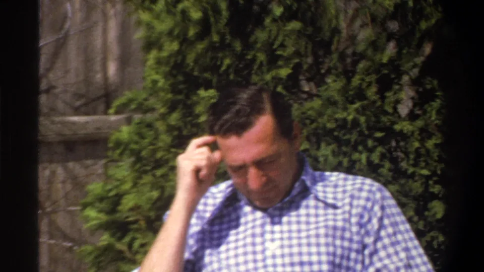 The man remains seated outside, looking down with one hand near his forehead against foliage and a stone wall in the 1969 footage.