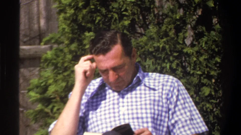 In 1969 Cape Town home-movie footage, a seated man in a blue-and-white checkered shirt reads outdoors near shrubs and a fence.