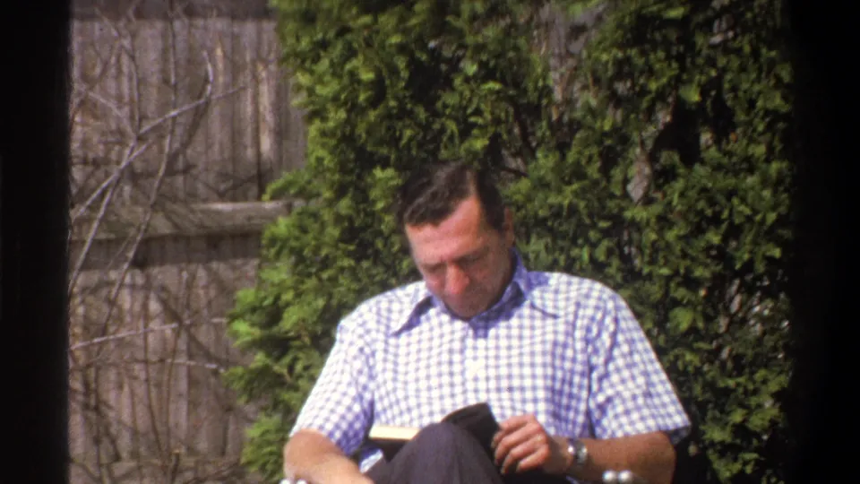 A wider 8mm frame shows the man seated in a backyard chair, looking down at a book with a fence and dense greenery behind him.