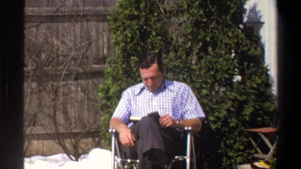 A wider 1969 Cape Town home-movie frame shows the man reading in a folding chair, with a backyard fence, shrubs, and patio items nearby.