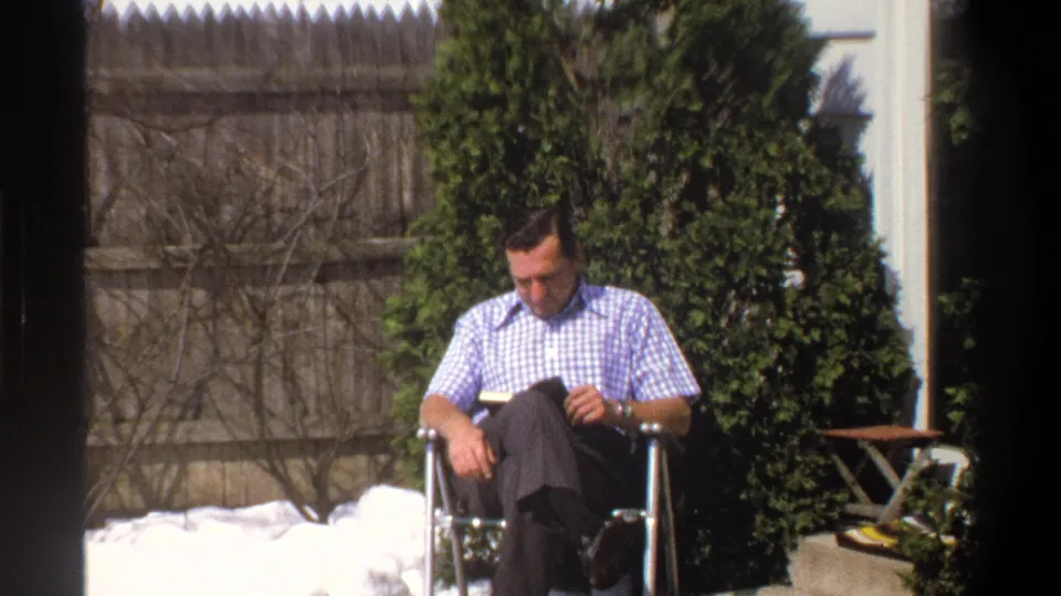 1969 Cape Town home-movie frame showing a man in a checkered shirt reading outdoors beside snow, shrubs, and a wooden fence.