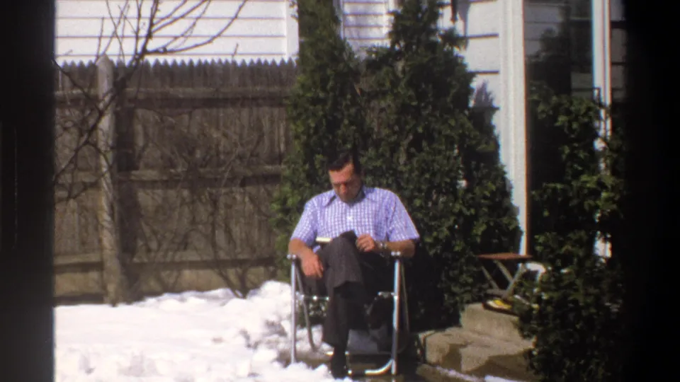 1969 Cape Town frame of a seated man reading in a snowy yard, with fence, shrubs, and house exterior visible behind him.