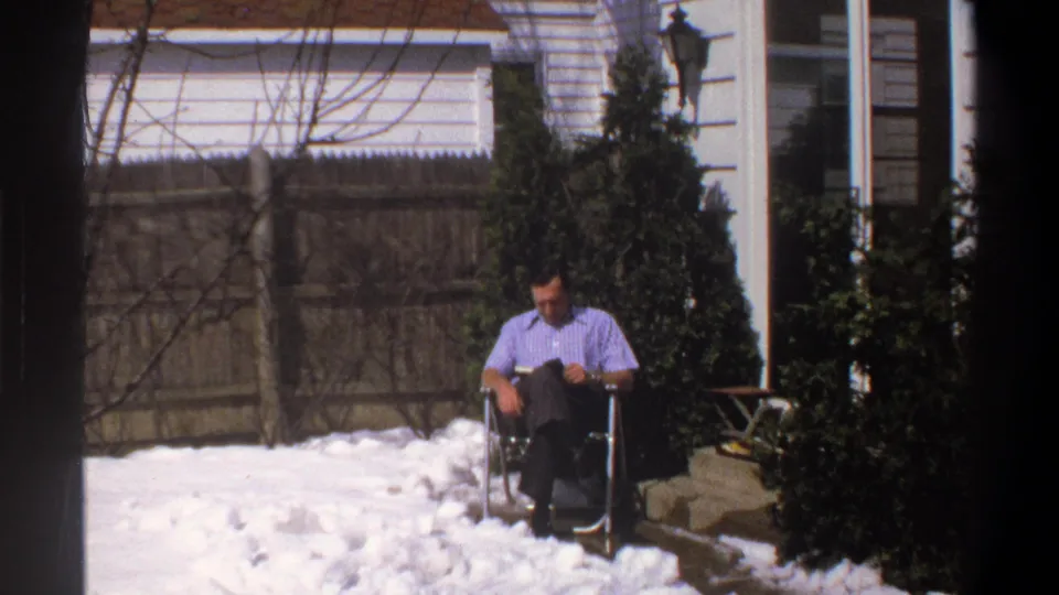 1969 Cape Town home-movie still with a wider view of a man reading in a chair on snow near shrubs, a fence, and a house wall.