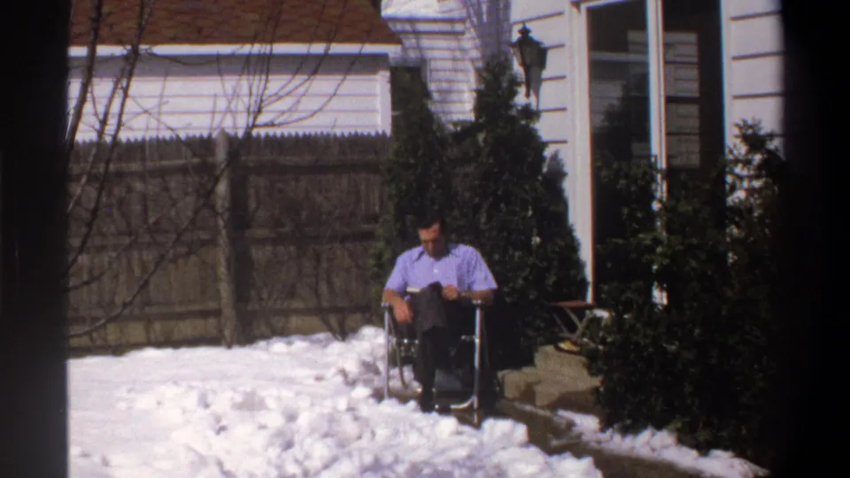 1969 Cape Town frame showing a man reading from a chair in a snowy backyard, bordered by shrubs, a fence, and house siding.