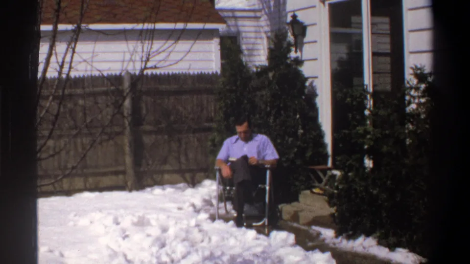 1969 8mm frame from Cape Town metadata showing an adult man seated outdoors in snow near a fence, shrubs, and house siding.
