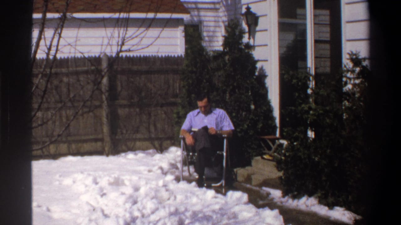 1969 Cape Town home-movie frame with a seated man in a snowy residential yard, looking down near evergreen shrubs and a wooden fence.