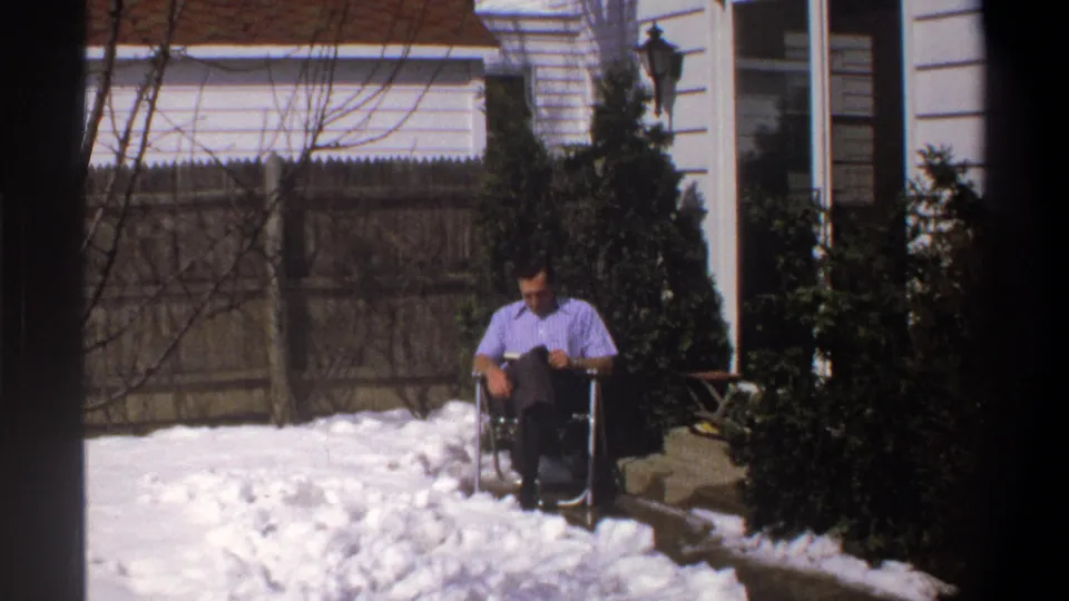 Home-movie frame from the 1969 Cape Town clip showing a man seated in snow outside a house with shrubs and a wooden fence behind him.