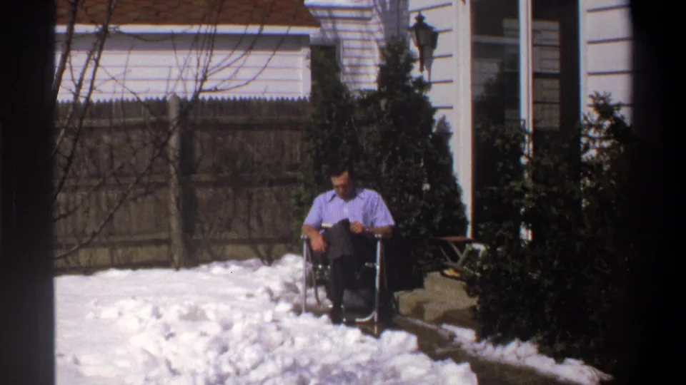 Final frame of a 1969 Cape Town 8mm clip with an adult man seated in a snowy yard near shrubs, a fence, and a house entrance.