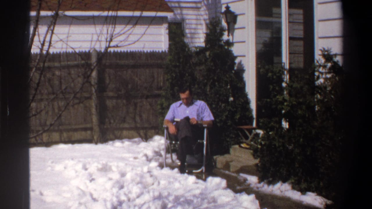 Man in a light blue shirt sits in a folding chair beside snow in a fenced yard.