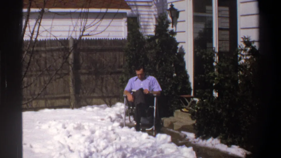 1969 Cape Town home-movie frame showing a man seated in a folding chair near snow patches, shrubs, fencing, and house exteriors.