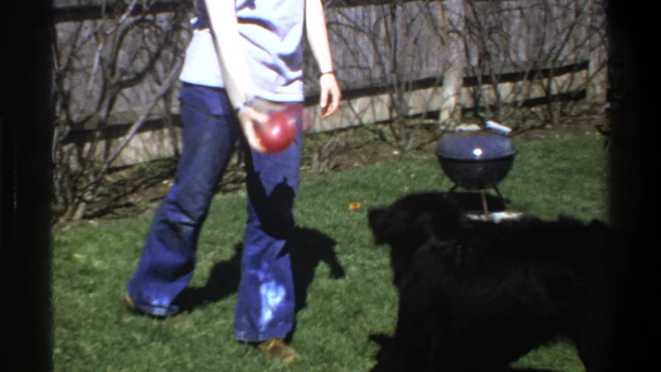 1969 Cape Town home-movie still showing a person in flared jeans with a red ball as a black dog waits in a backyard.