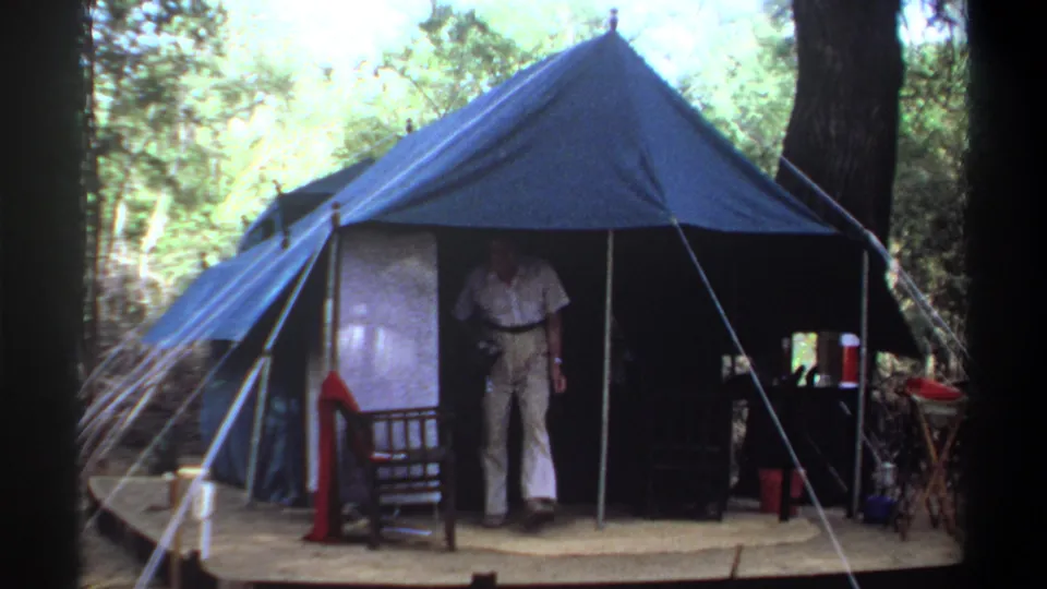 Adult man appears in the entrance of a blue canvas tent at a wooded camp identified as Khartoum, Sudan, 1969.