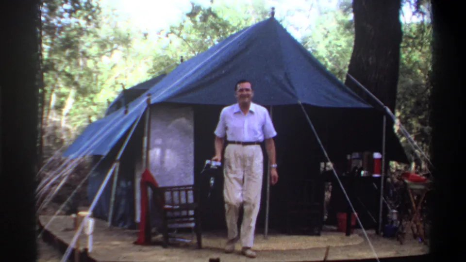 Adult man in a light short-sleeved shirt and trousers walks from a blue canvas tent, holding a camera in 1969 Sudan footage.