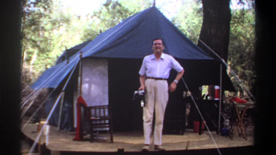 Adult man smiles toward the filming camera outside the blue canvas tent at the Khartoum, Sudan camp, dated 1969 by metadata.