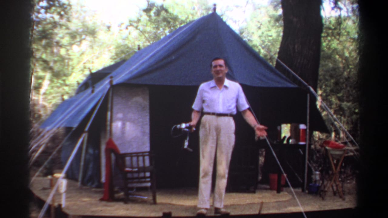 Adult man in a light shirt and cream trousers stands before a blue canvas tent at a wooded Khartoum campsite in 1969 footage.