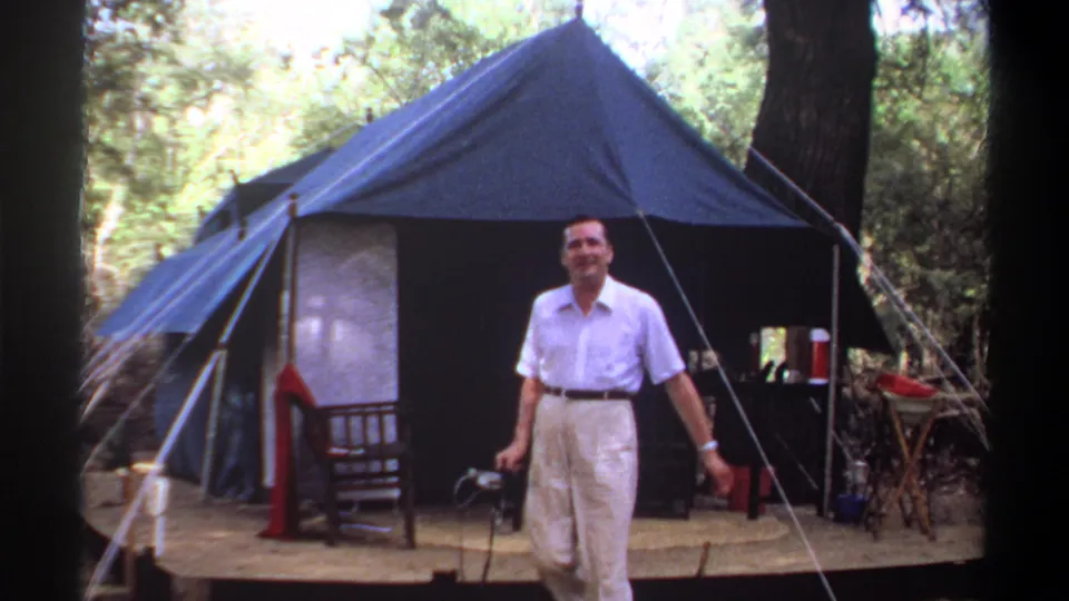 Adult man walks out from a blue canvas tent in wooded 1969 Khartoum home-movie footage, holding a small tool or device.
