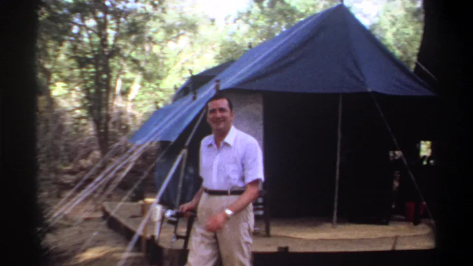 Adult man stands closer to camera beside the blue tent and wooded path, holding a small handheld tool or device.