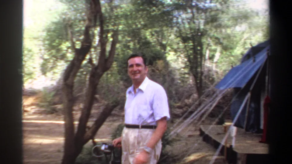 Smiling adult man stands near trees at a wooded campsite, with part of the blue tent visible in 1969 Khartoum footage.