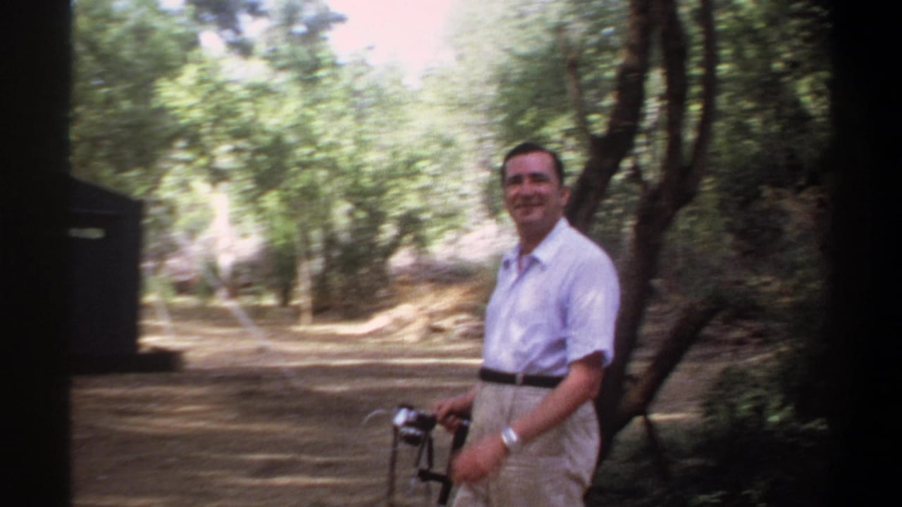 In 1969 Khartoum home-movie footage, a smiling man with a camera walks through a wooded campsite area.