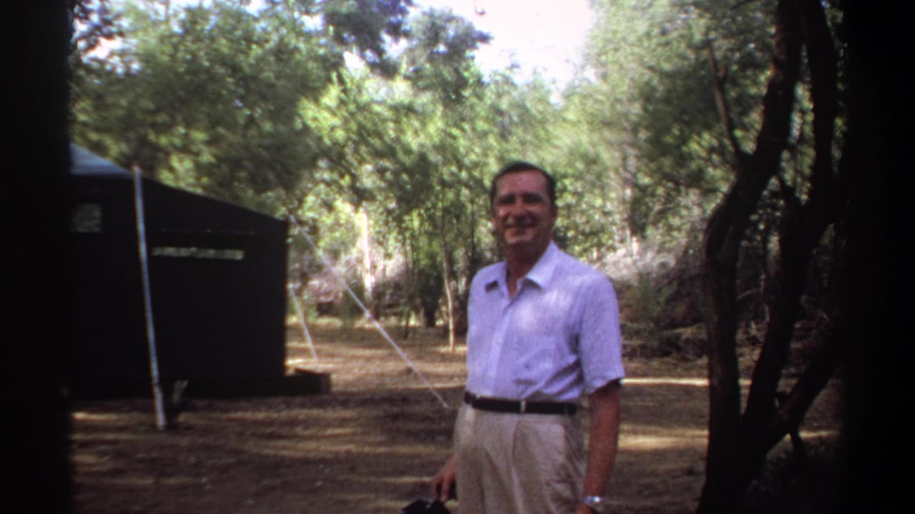 Smiling man stands near a dark tent among trees in a shaded campsite.