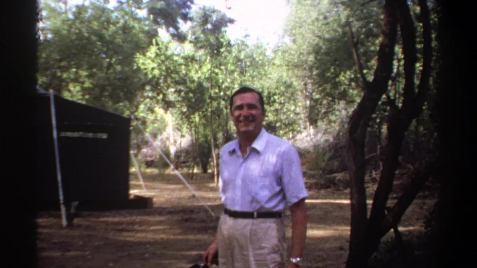 The man faces the lens and smiles in a wooded campsite setting, with a dark tent visible behind him in 1969 Khartoum footage.