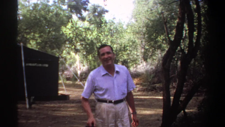 A smiling man in light clothing stands beside trees and a dark campsite tent in vintage 8mm footage identified as Khartoum, 1969.