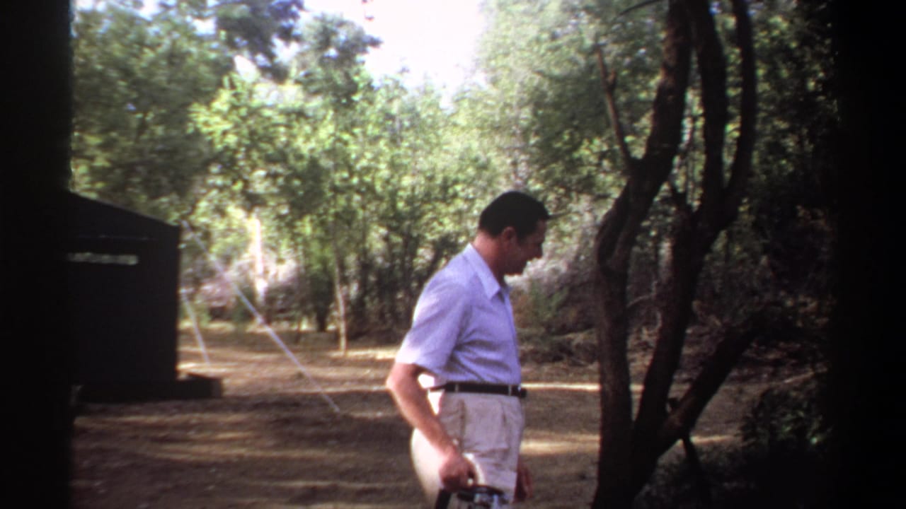 Man in a light shirt walks through a wooded campsite holding a camera.