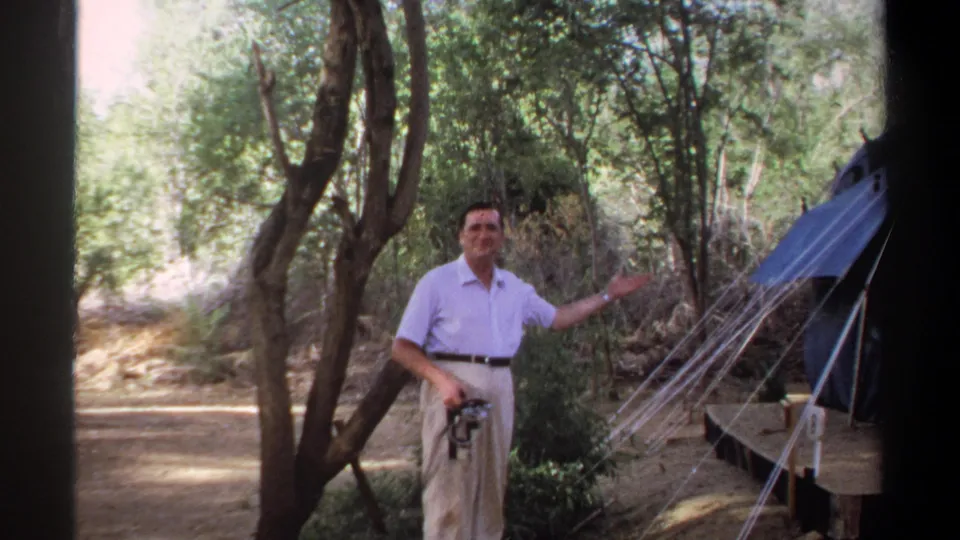 In the wooded campsite, the man faces the camera and gestures toward the pitched blue tent beside him.